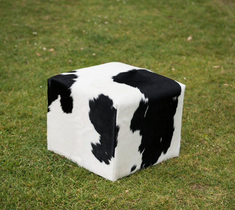 A cube-shaped pouf ottoman with a black and white cow print pattern, placed on a green grass lawn.